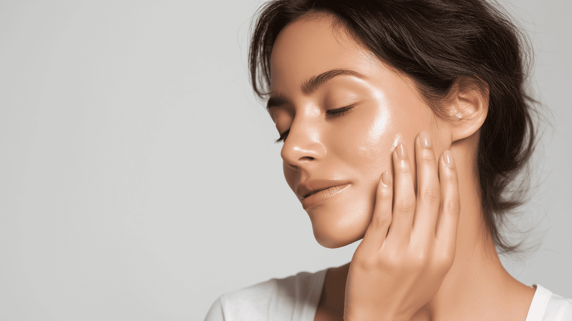 Woman with glowing skin gently touching her face against a neutral background.
